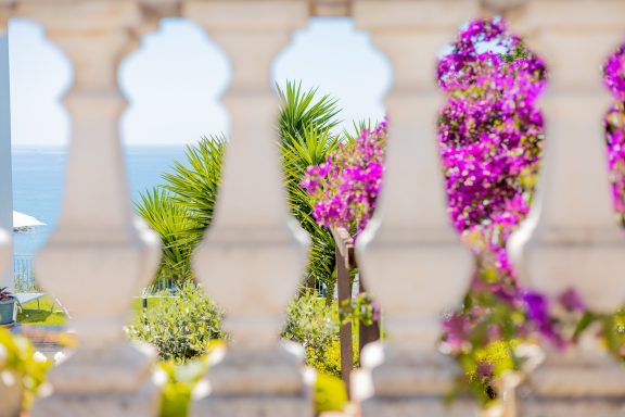 Vista marina attraverso un parapetto, con fiori viola e vegetazione tropicale.