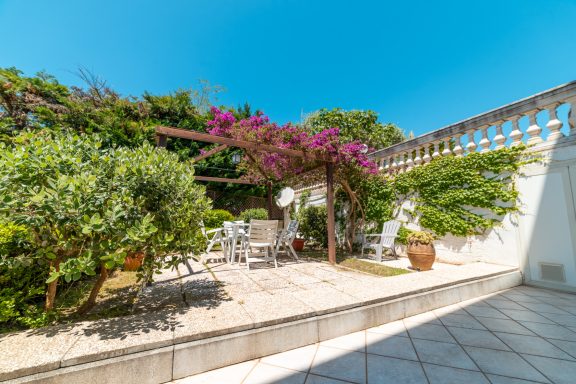 Giardino con pergolato e fiori, sedute bianche su pavimento in ceramica.