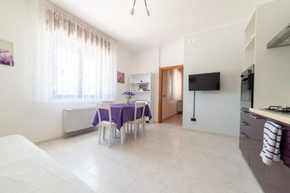 Interno di un appartamento luminoso con cucina, tavolo viola e TV montata a parete.