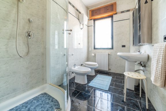 Bagno moderno con doccia, sanitari e pareti chiare, luminoso da una finestra.